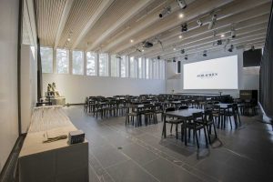 Gösta’s Kivijärvi Hall used as a meeting space. There is a big screen in the back, with groups of tables in front of it and large windows.