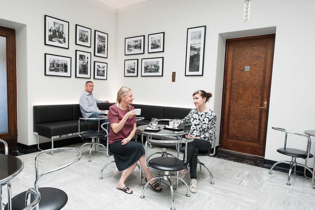 Kaksi naista istuu pyöreän pöydän äärellä kahvilla. Heidän takanaan mustalla sohvalla istuu mies kahvittelemassa. Taustalla oleva seinä on täynnä mustavalkoisia valokuvia.