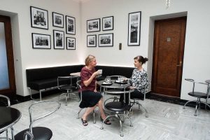 Two women seated at a round table drinking coffee. The wall behind them is full of black and white photos.