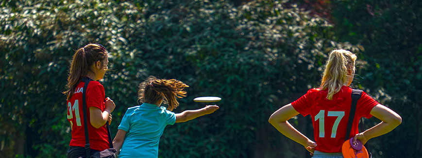 Kaksi punapaitaista naista seuraa kun sinipaitainen nainen heittää frisbeegolfkiekkoa.