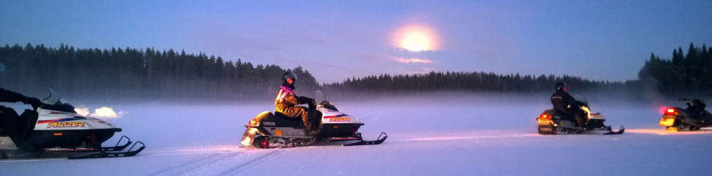 Neljä moottorikelkkailijaa ajaa illan hämärtyessä lumisen järven yli, taustalla laskee aurinko puiden taakse.
