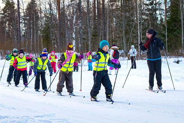 A line of day-care children wearing high-visibility vests, skiing on tracks in a field.