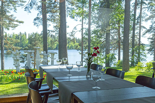 Ravintolasalin pöytä, pöydällä kukkamaljakko. Taustalla iso ikkuna josta näkyy moderni veistos ja järvimaisema sekä mäntymetsää.