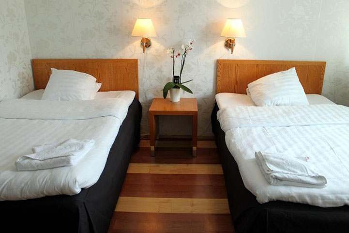 Hotellihuone, kaksi pedattua sänkyä valkoisine lakanoineen, seinälampuissa valot.