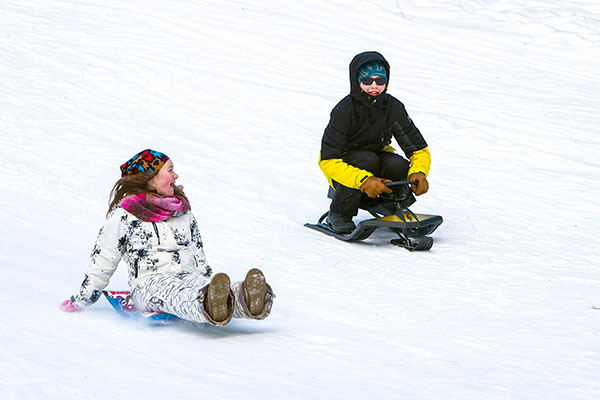 Two children whizzing down a hill, one on a sledge and one on a snow racer