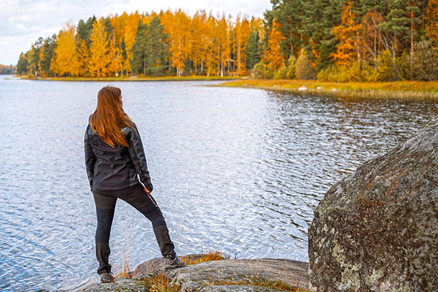 Ulkoiluvaatteisiin pukeutunut ruskeahiuksinen nainen katsoo kalliolta syksyistä järvimaisemaa.