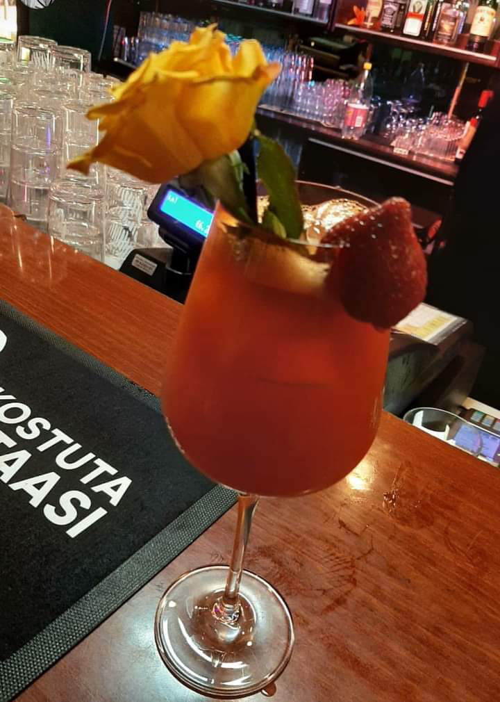 Herkullisen näköinen punainen drinkki, lasin reunalla mansikka ja keltainen ruusu.
