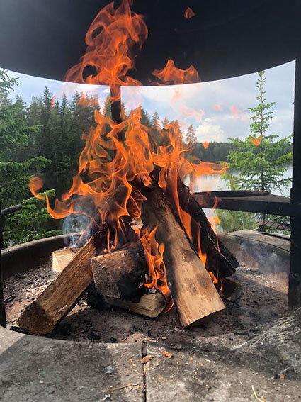 An open fire with orange flames. Spruce trees in the background and a lake partly visible behind them.