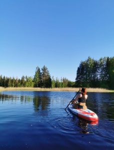 A woman paddling on a SUP board on a calm lake. Blue sky, reeds and a forest are visible in the background.