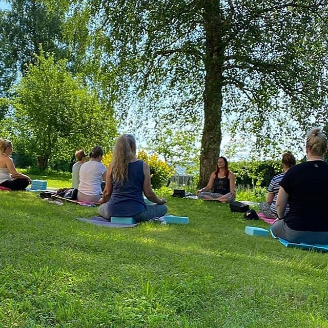 A yoga instructor and a group of students sitting cross-legged on yoga mats in the shade of a tree in a park.
