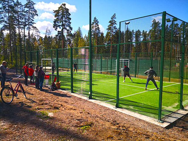 A group of people watch four men playing padel on an outdoor court on a sunny day.