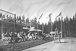 A black-and-white photo of an audience watching a tennis match behind the umpire, in front of the Tennis Pavilion and on its roof.