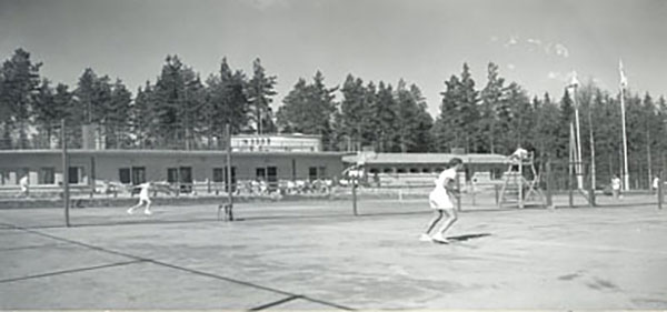 Mustavalkoisessa kuvassa kaksi henkilöä pelaa tennistä yleisön edessä.
