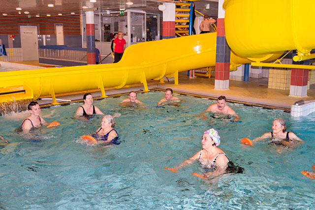 People doing aquatic exercise next to a yellow water slide in a warm water pool.