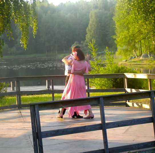 A woman dressed in a long pink dress dances with her partner at an open-air dance pavilion on a summer evening. There is a calm lake and green birch trees in the background.