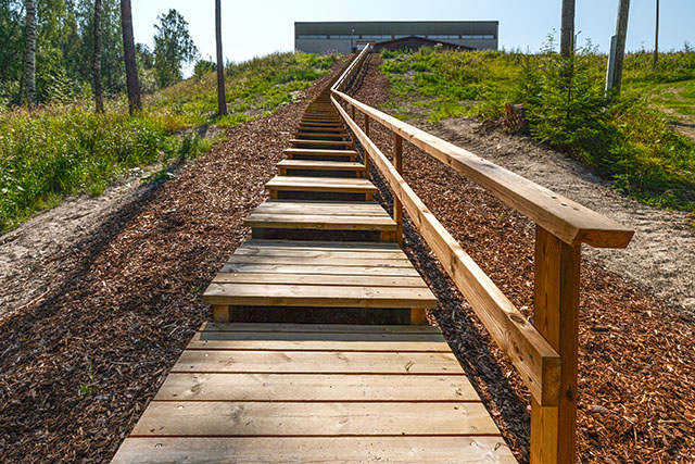 The bottom of the stairs, with woodchips, grass and a few trees. The indoor sports facility is visible at the top of the slope.