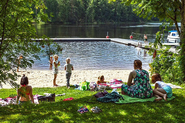 Families with children sitting in the shade on a summer day. In the background, two boys are jumping off a dock into the water.
