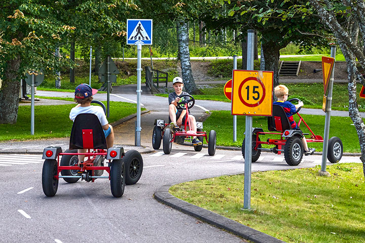 Kolme lasta kohtaavat liikennepuiston risteyksessä, jossa on erilaisia liikennemerkkejä.