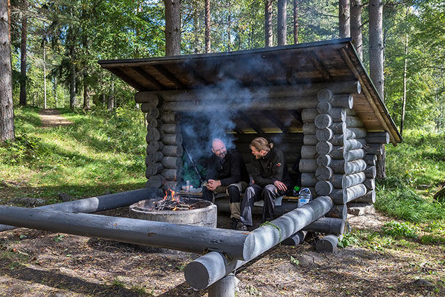 Two men sitting at a lean-to surrounded by trees, grilling sausages.