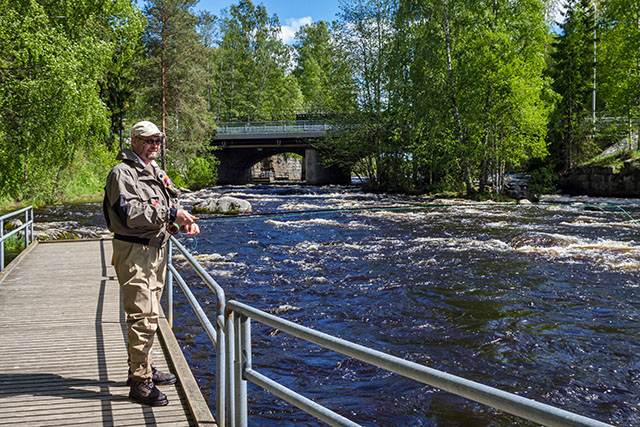 A smiling fly fisher standing on a fishing dock on a sunny summer day, fishing. In the background is a bridge across the rapids and green trees.