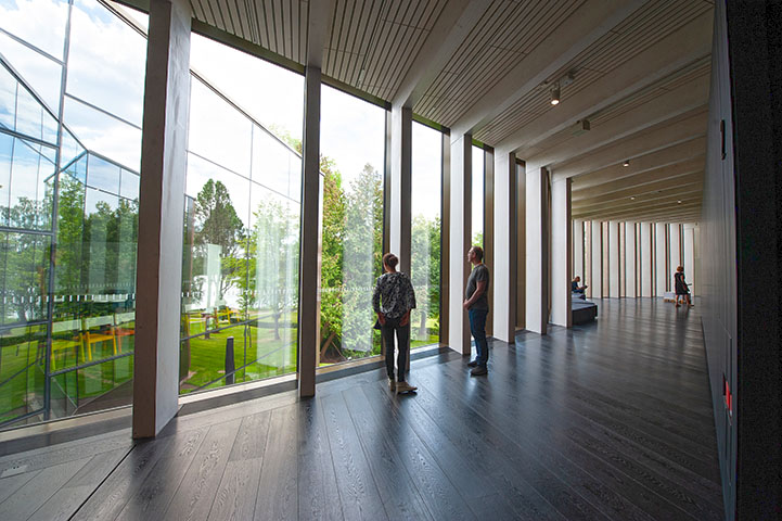 Serlachius-museo Göstan paviljongin käytävä. Kaksi ihmistä ihailee ulkona olevaa vehreää puistoa käytävän lattiasta kattoon asti olevista isoista ikkunoista.