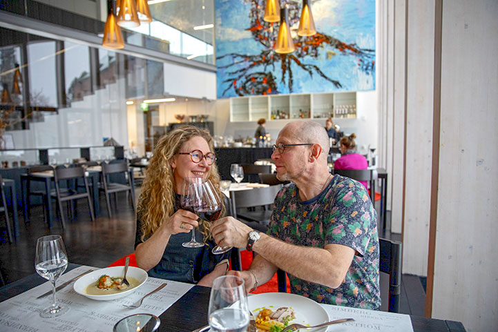 A smiling couple sitting at a table, toasting wine glasses, with plates of delicious food in front of them.