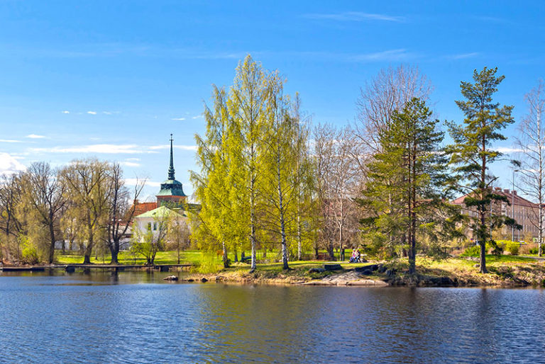 Koskelanlampi kesällä. Rannalla näkyy puiden ympäröimänä penkki, jossa istuu ihmisiä. Taustalla Mäntän keskustan rakennuksia ja Mäntän kirkon torni.