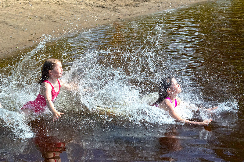 Kaksi tyttöä pinkit uimapuvut päällään rientävät uimaan Vilppulan sataman uimarannalla veden roiskutessa.
