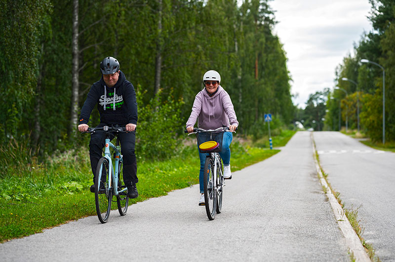 Mies ja nainen pyöräilevät pyörätiellä vehreän metsän katveessa.