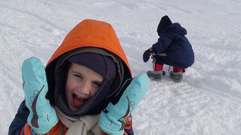Nuori lapsi talvivaatteissa nauraa täydestä sydämestä, taustalla näkyy lunta ja toinen lapsi kyykyssä.