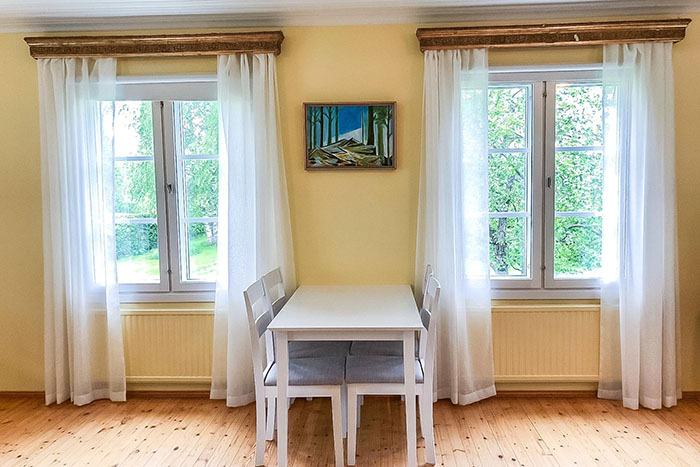 Valkoinen suorakulmainen pöytä neljällä valkoisella tuolilla, kahden ikkunan välissä. Pöydän yläpuolella, vaaleankeltaisella seinällä maalaus.