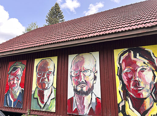 Punaisen puutalon seinässä on neljä seinänkorkuista potrettia, taustalla näkyy sininen taivas ja kuusenlatva.