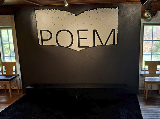 Pienen lavan taustaseinällä lukee isoilla kirjaimilla POEM, lavan kummallakin puolella näkyy vanhan rakennuksen ikkunat sekä tuoleja.