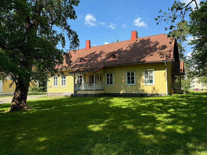Keltaisen ison punakattoisen puutalon edessä on nurmikkoa ja iso lehtipuu, jonka varjot näkyvät nurmikolla ja talon seinällä sekä katolla. Taustalla sininen taivas.