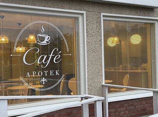 Kahdesta isosta ikkunasta näkee ulko sisälle kahvilan viihtyisiin tiloihin, vasemmassa ikkunassa Café Apotekin pyöreä, valkoinen logo.
