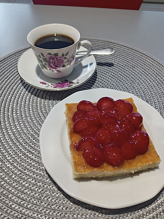 Pöydällä odottavat täytetty ruusukuviollinen kahvikuppi sekä herkullisen näköinen mansikkakakku.